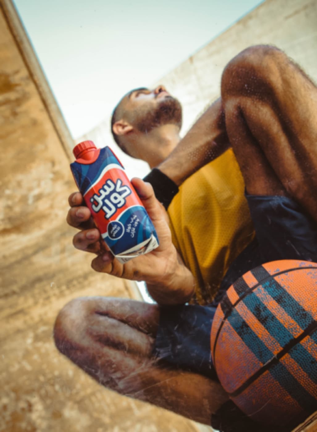 A basketball player sitting on the court with a ball, drinking from a Suncola carton.