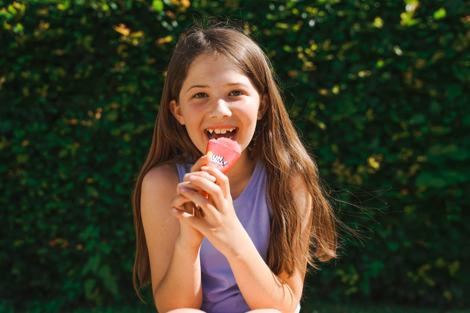 A girl in front of a hedge eating a Sun Lolly.