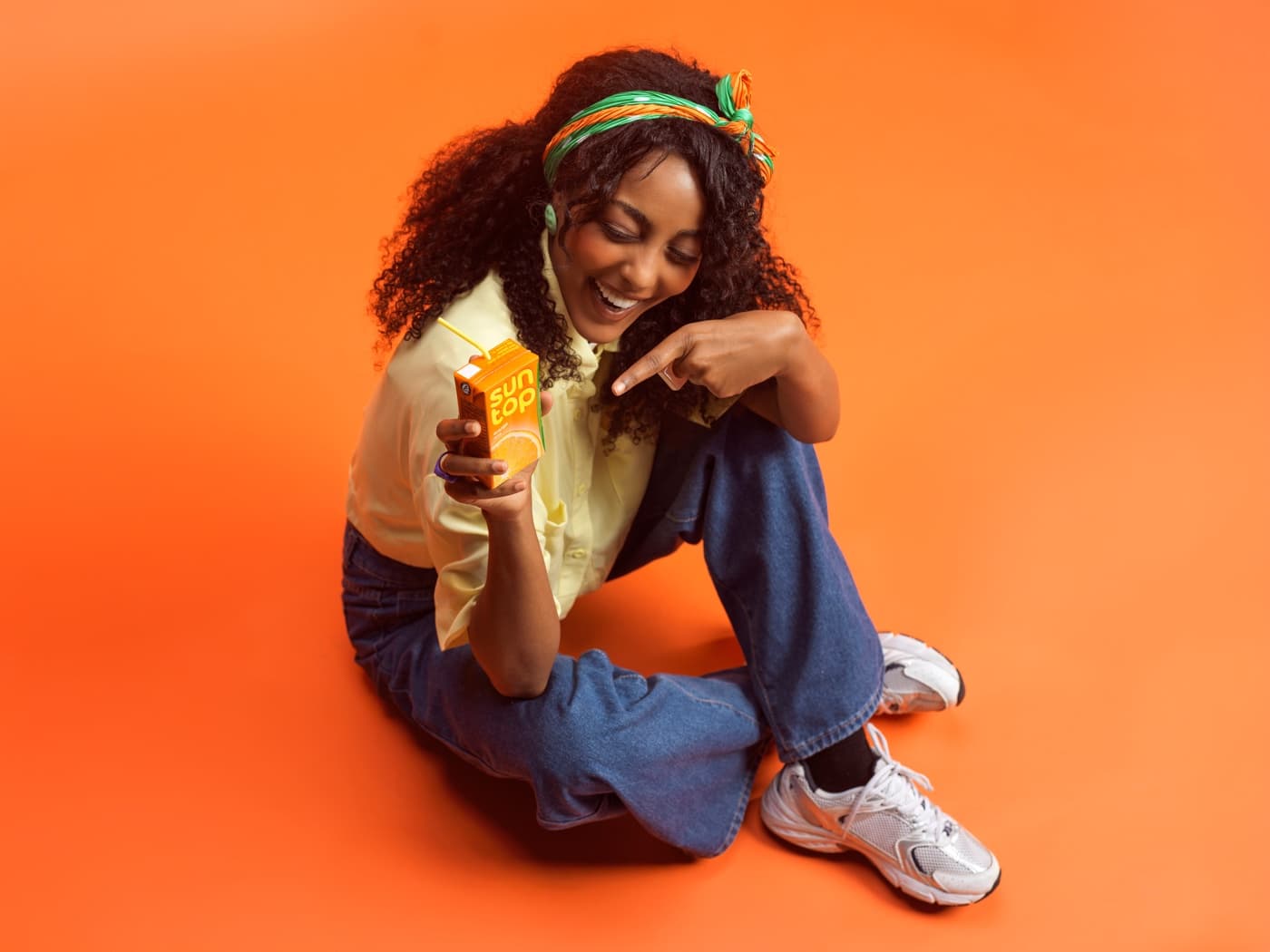 A young woman seated on an orange ground, laughing, a Suntop carton in her hand.
