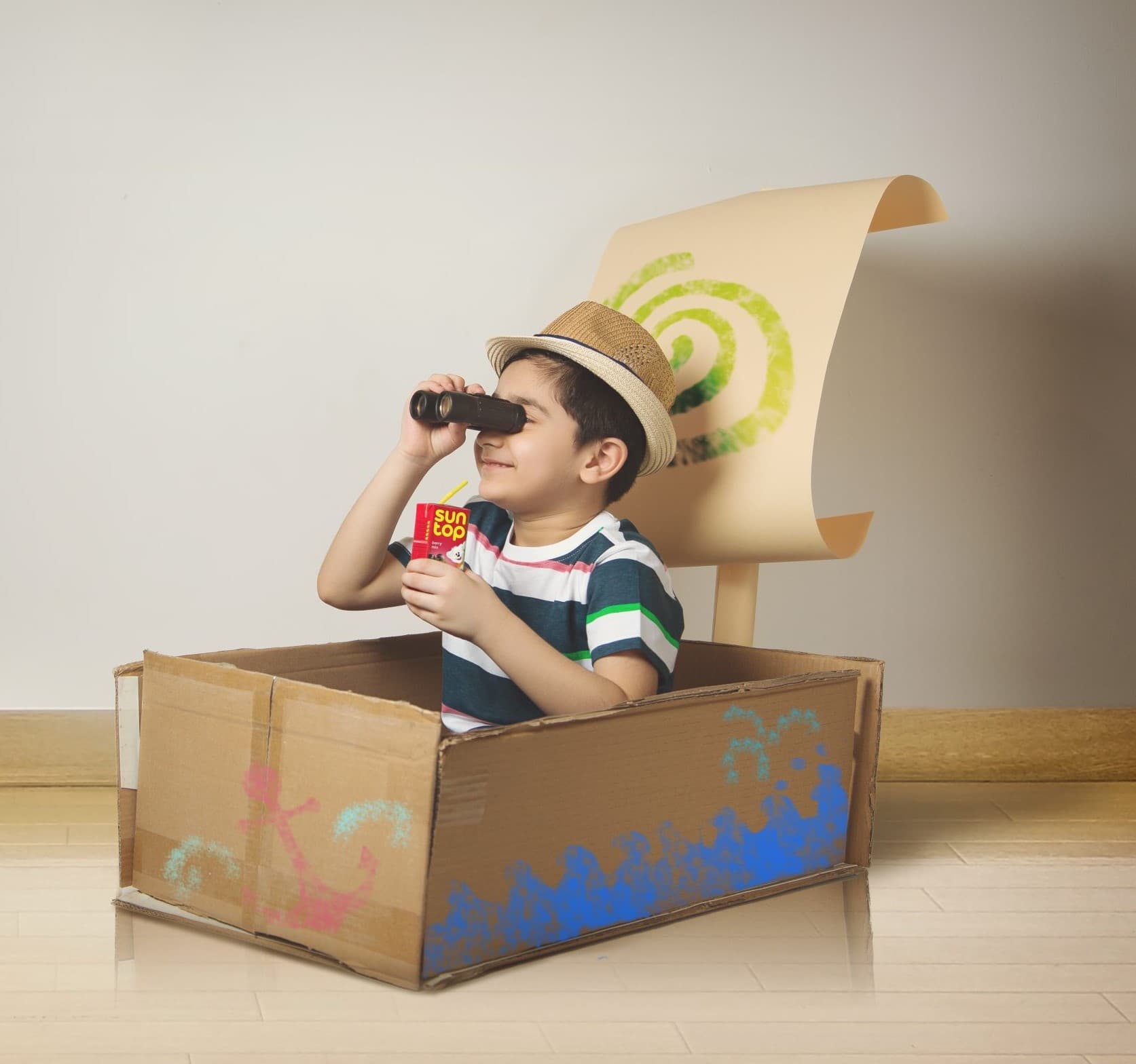 A boy sitting in a cardboard boat with a paper sail, binoculars raised, a Suntop carton in his hand.